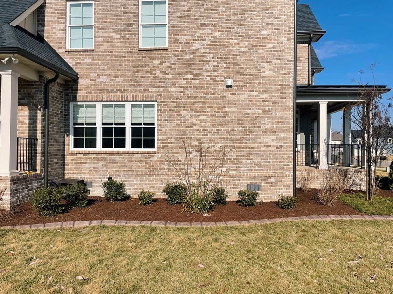 Tan brick house with white-framed windows, a porch, and landscaped bed bordered by red brick.