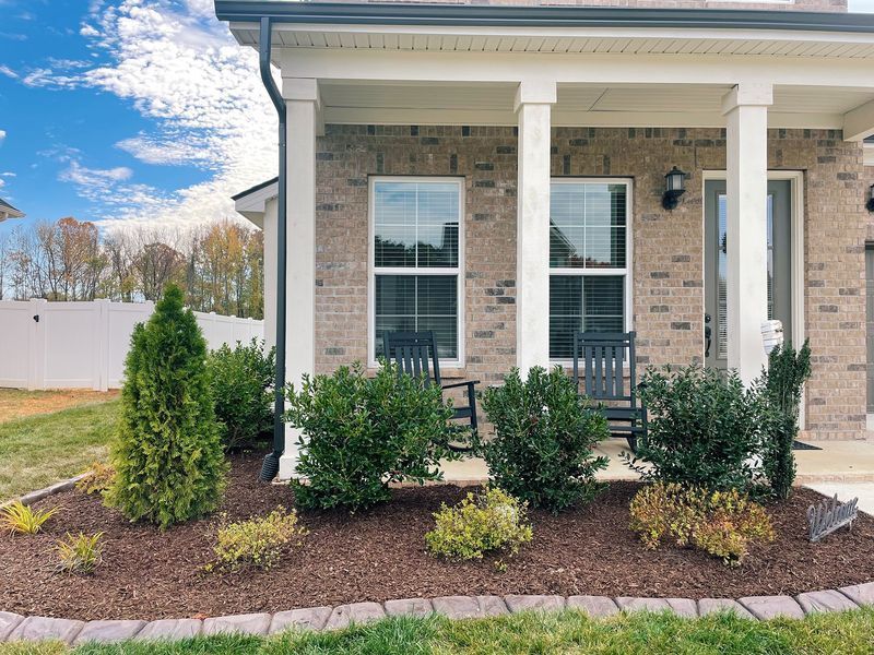 Front porch of a brick house with bushes and mulch in front. Two rocking chairs sit on the porch.