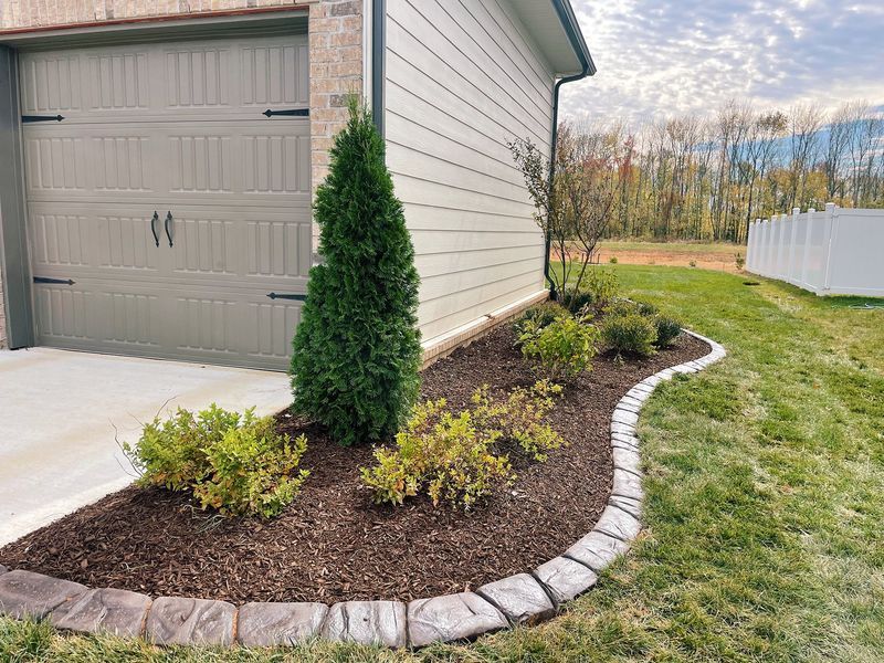 A landscaped garden bed with mulch, plants, and a stone border next to a garage.
