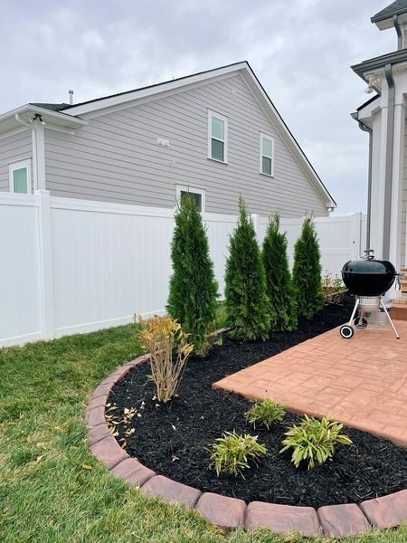 Backyard with a brick patio, green plants, black mulch, and white fence. A grill sits nearby.