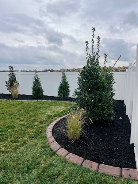 Green lawn with a curved bed edged with bricks, containing plants and dark mulch, along a white fence.