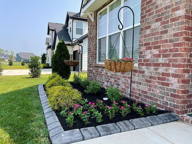 Flowerbed with pink flowers, black mulch, and brick home. A hanging basket is beside the window.