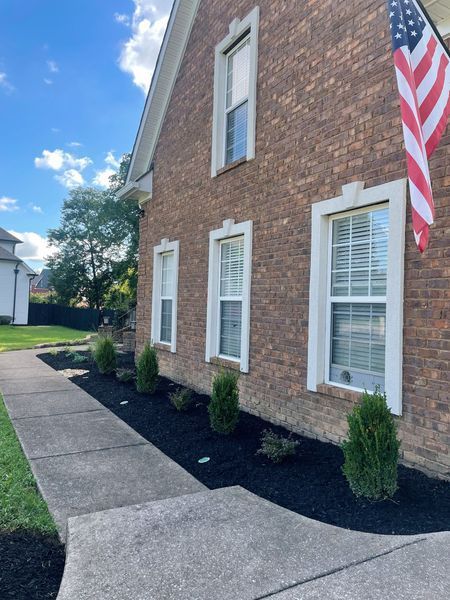 Brick house with white-trimmed windows and an American flag. Green shrubs line the black-mulched flower bed.