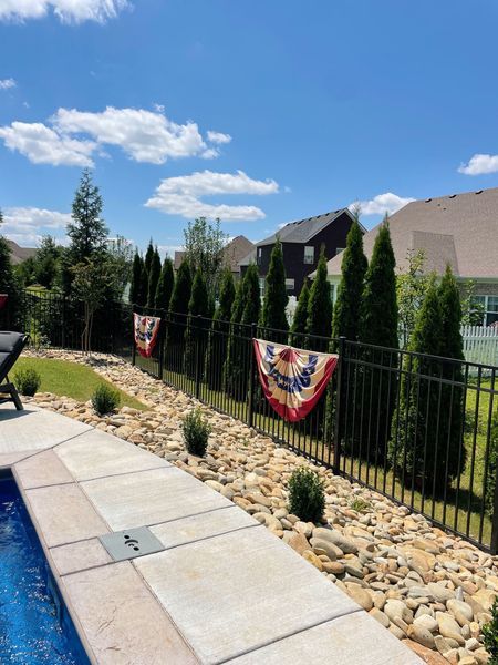 Backyard with pool, black fence, green trees, and red, white, and blue bunting under a blue sky.