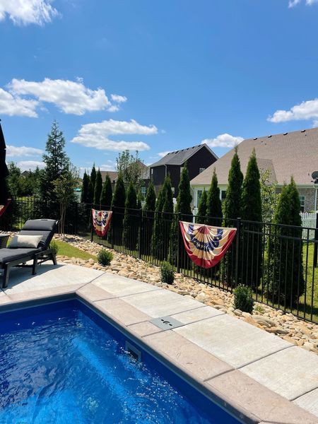 Poolside view with evergreen trees, black fence, and patriotic bunting under a sunny sky.