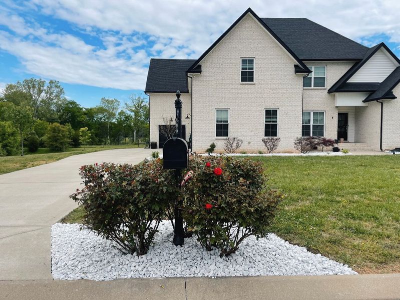 Two-story house with black mailbox and red flowers surrounded by white stones on green lawn.