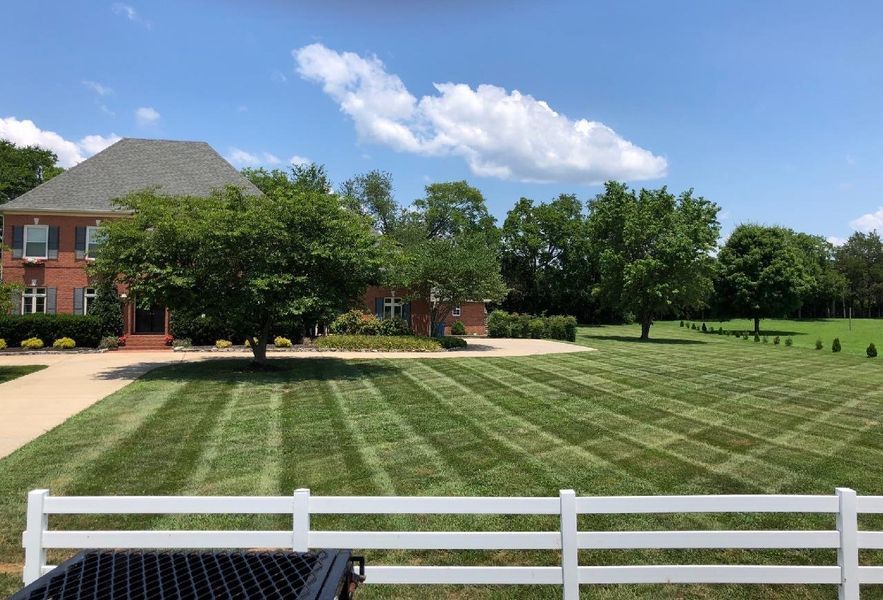 Lawn with striped mowing pattern, brick house, trees, blue sky with clouds, and white fence in foreground.