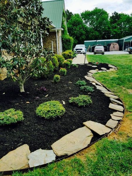 Landscaped flower bed with dark mulch, stone border, and green shrubs in front of a house.