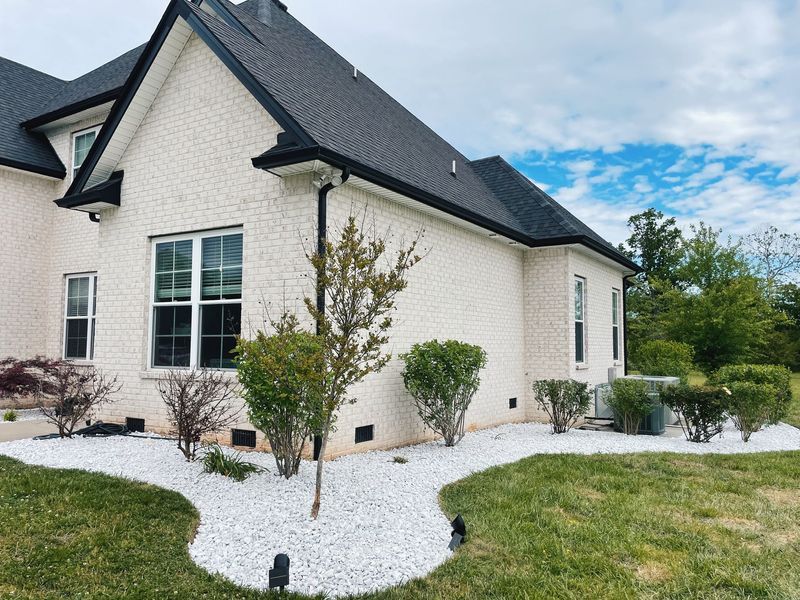 White brick house with black trim, landscaping with white stones and green bushes.