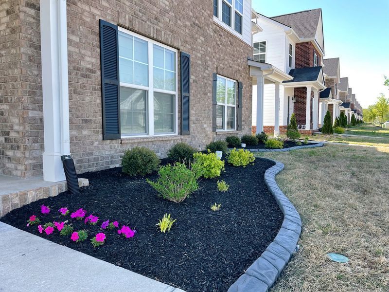 A brick house with a flower bed and black mulch. Pink and yellow flowers, shrubs, and a gray border are in the bed.
