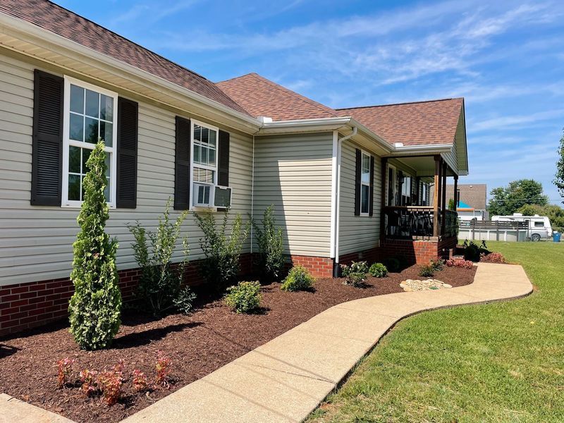 Tan ranch-style house with brown roof, red brick, and sidewalk, with landscaping and blue sky.