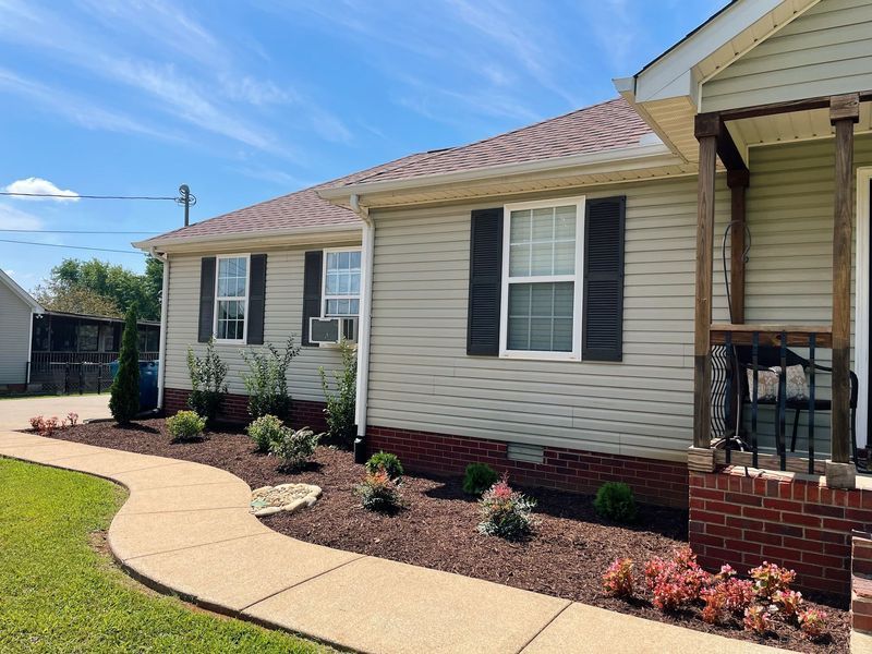 Tan house with brown roof, black shutters, and a concrete walkway lined with plants.