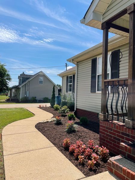 Pathway leading to a beige house with landscaped flower beds and a view of another house on a sunny day.