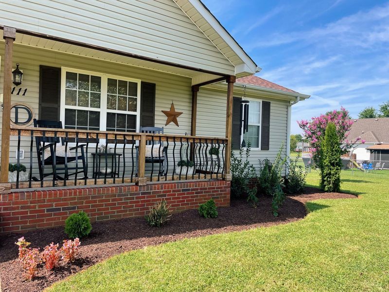 Beige house with a porch, brick base, and brown trim, with a landscaped yard and blue sky.