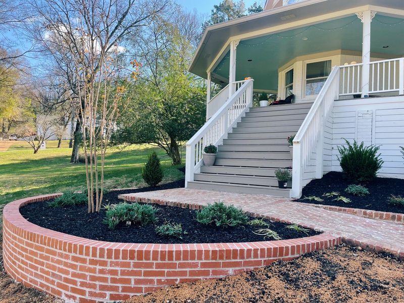 A brick-lined garden bed and walkway lead to a white house with a porch, blue ceiling, and gray stairs.