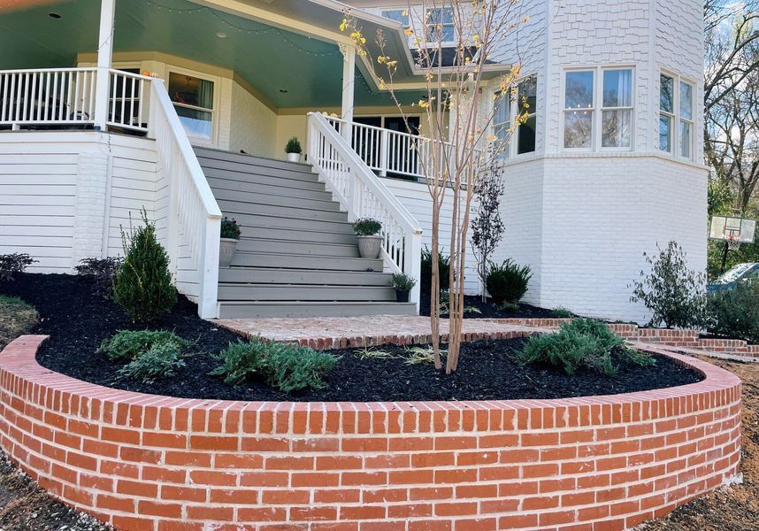 Brick planter with black mulch and shrubs in front of a white house with stairs leading to a porch.