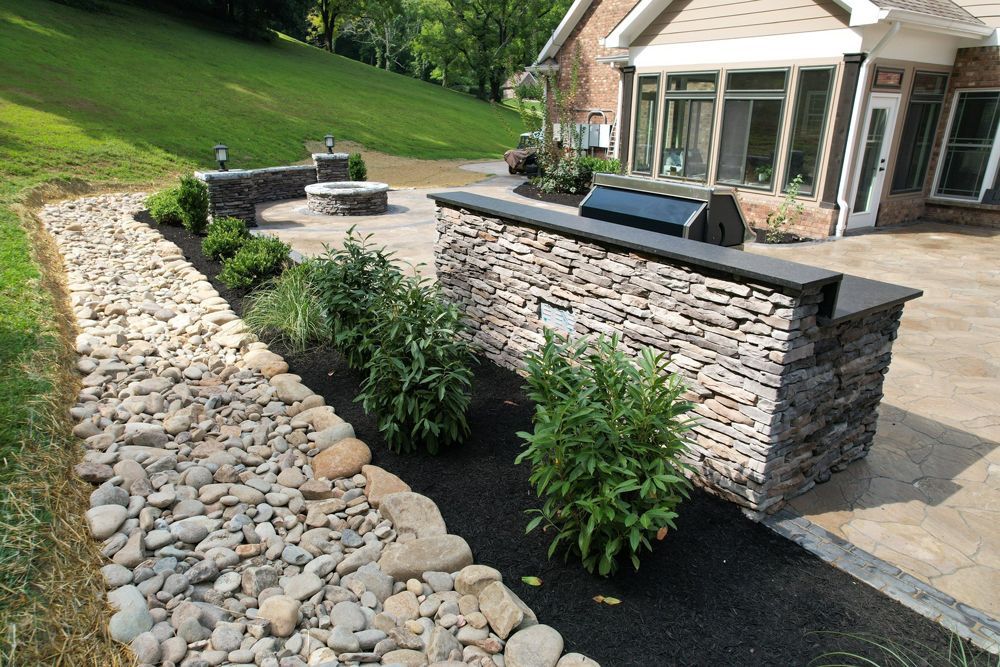 Stone patio with grill, fire pit, and retaining wall on a lawn next to a house.