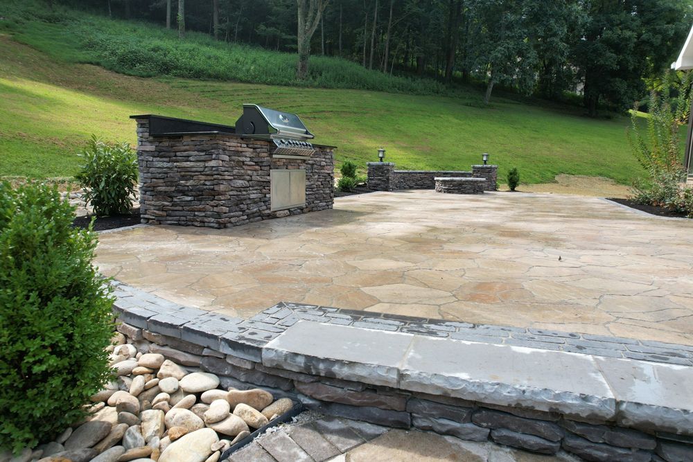 Stone patio with built-in grill, bench, and landscaping against a grassy hillside.
