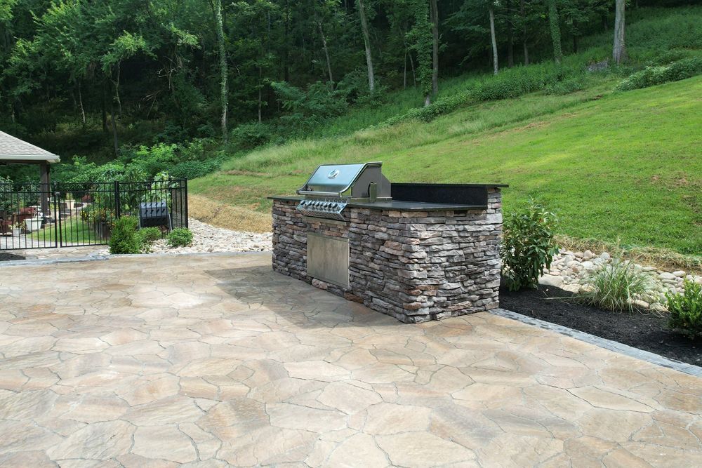 Stone-covered outdoor grill station on a stone patio, set against a grassy hillside.
