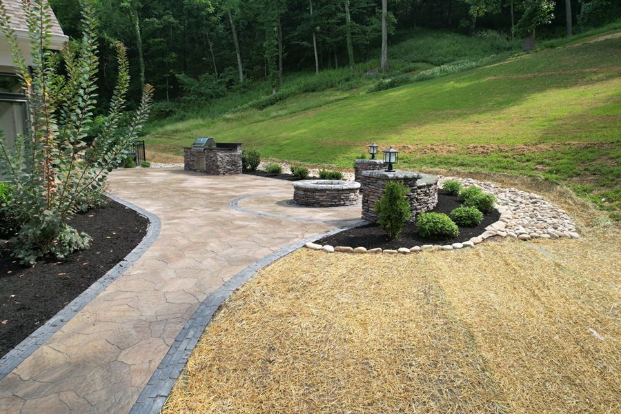 Stone patio with grill, fire pit, and landscaping on a grassy hillside.