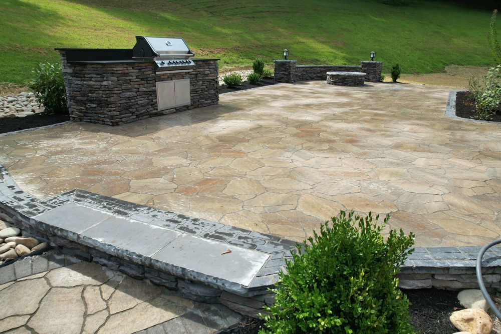 Stone patio with grill, fire pit, and stone walls; green grass in the background.
