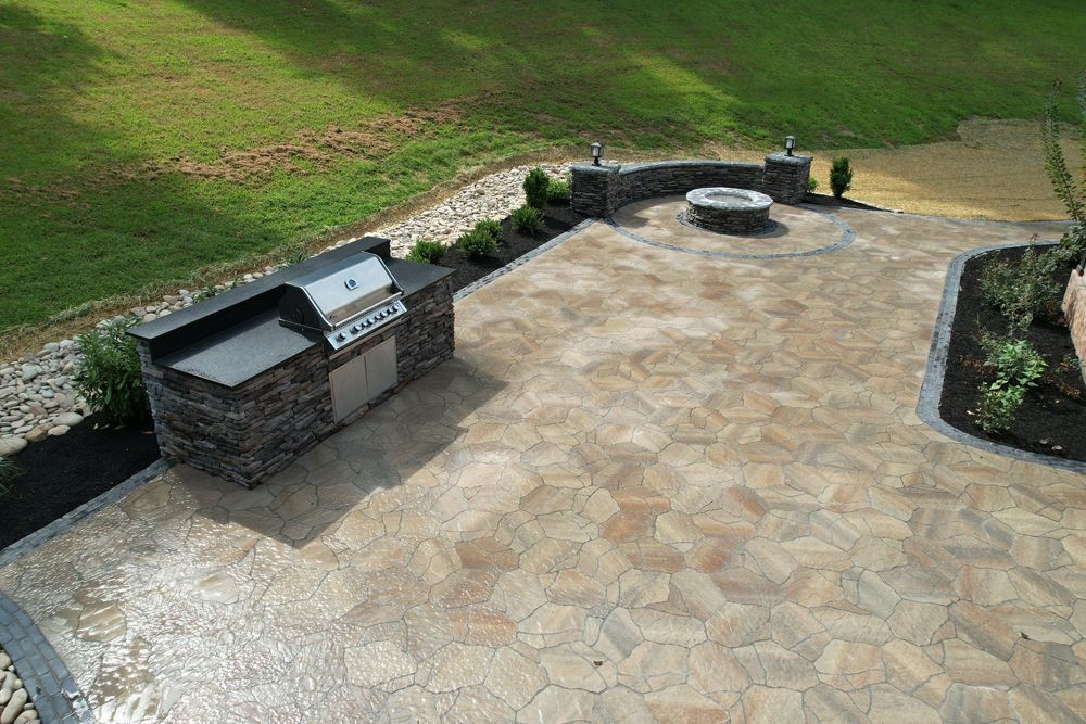 Stone patio with grill station, fire pit, and grass hill in background.