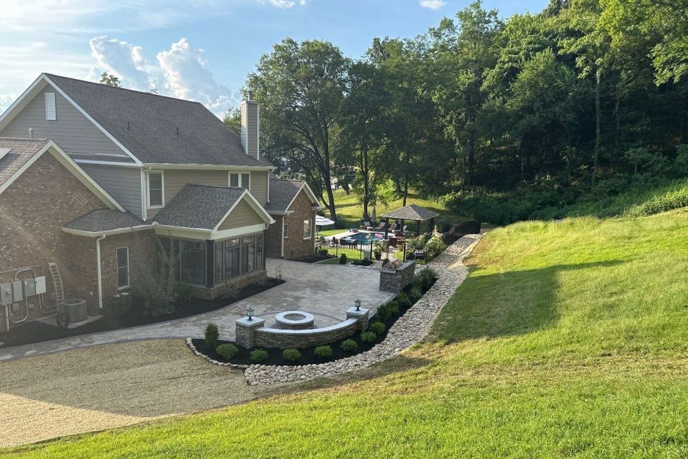 A large house with a stone patio, fire pit, and manicured landscaping, next to a grassy hillside.