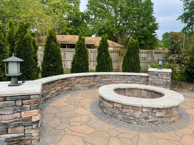 Stone patio with built-in fire pit, stone wall, arborvitae trees, and gray stone pavers.