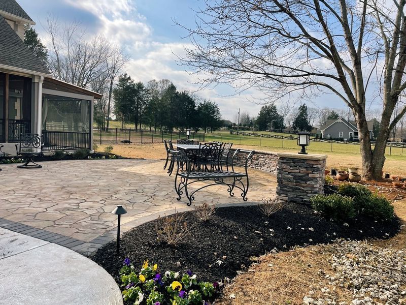 Stone patio with ornate bench, low stone wall, landscaping, and distant trees under a cloudy sky.