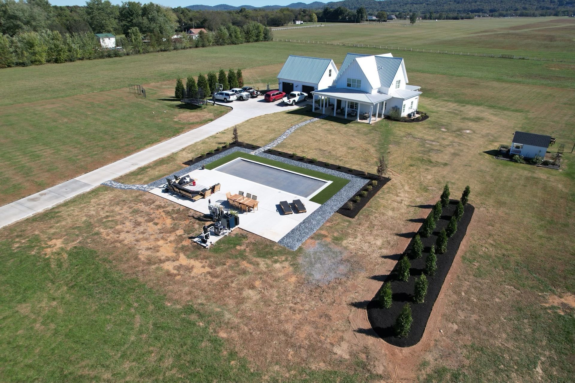 Aerial view of a white farmhouse with a pool, patio, and long driveway on a green field.