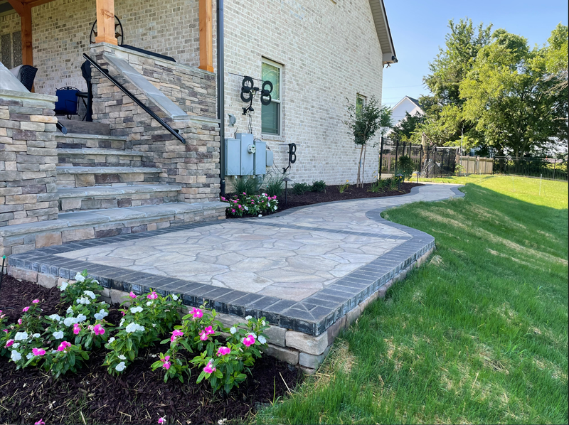 Stone patio with flower beds, leading to a house with brick exterior and stairs.