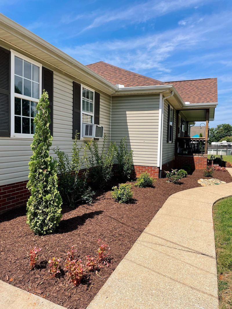 Tan house with brown roof and walkway, landscaped with mulch and plants.