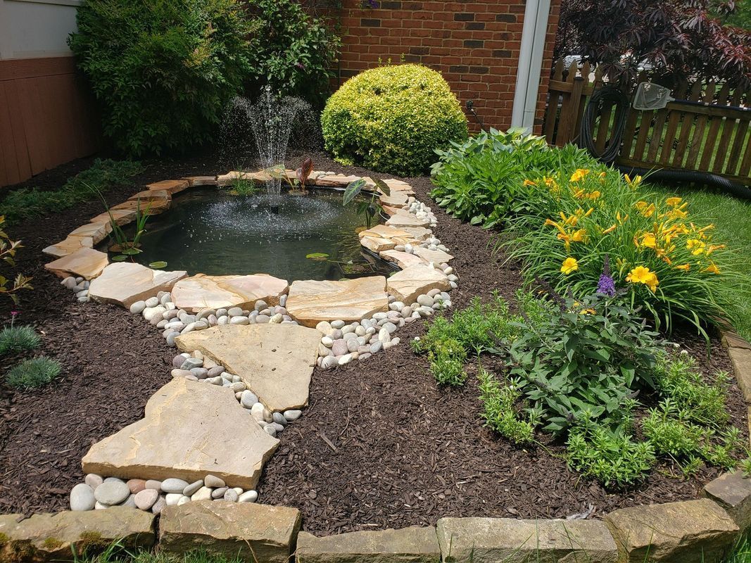 Small garden pond with fountain and stone path, surrounded by flowers and mulch.