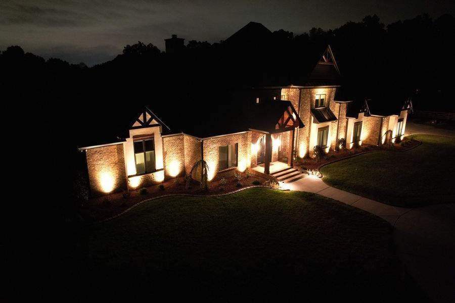 Lit-up home at night, exterior view with spotlighting on brick and stone facade, walkway, and a dark backdrop.
