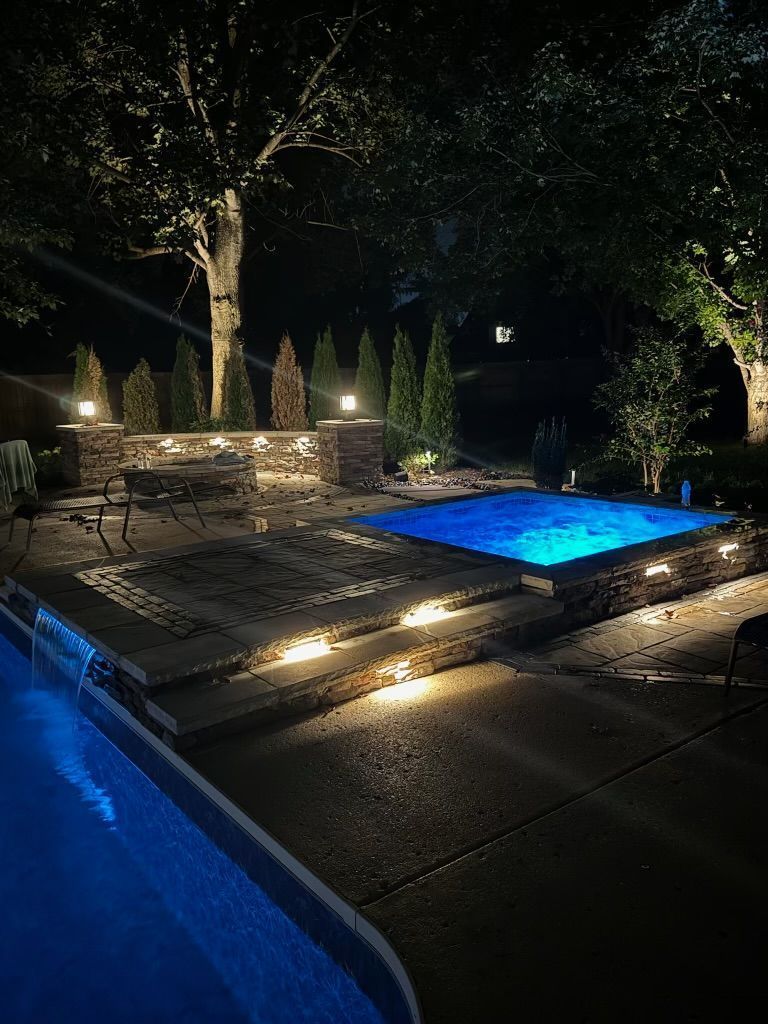 Nighttime view of a pool and spa, illuminated with blue and warm lights, stone wall and steps.