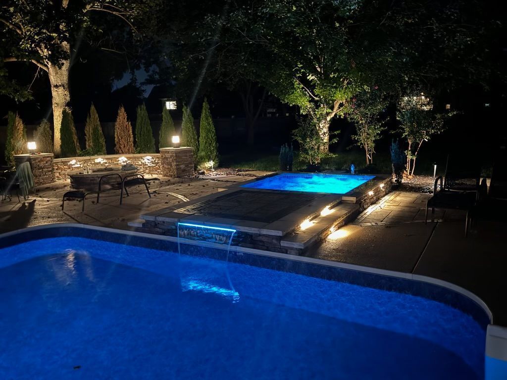 Nighttime shot of a backyard with a pool and illuminated hot tub surrounded by landscape lighting.