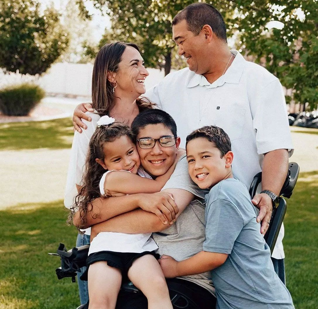 A family gathers outdoors for a group hug. A person sits in a wheelchair while others embrace them, all smiling.