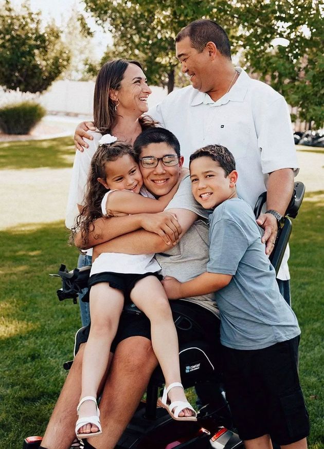 A family of five embraces in a park, with parents smiling at each other while their three children hug the child in front.