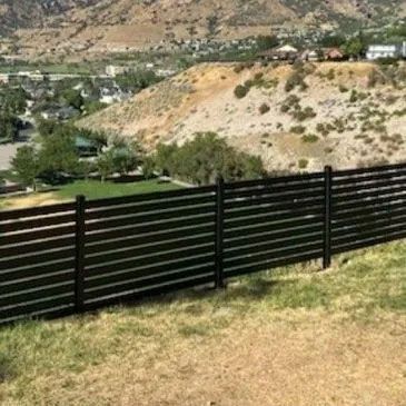 A dark, horizontal slat fence runs across a grassy hillside overlooking a valley with houses and distant mountains.