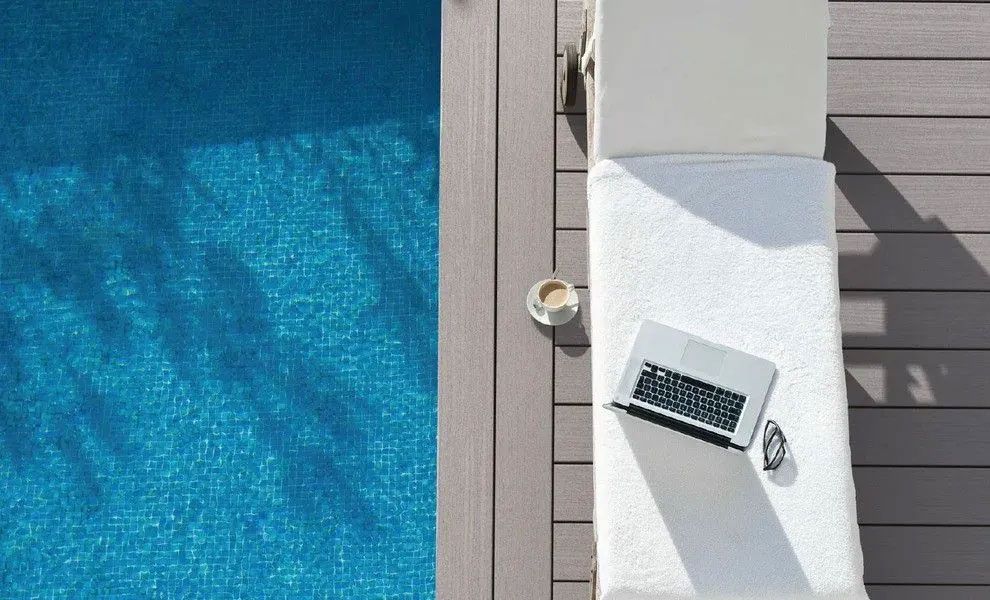A laptop and a cup of coffee sit on a white lounge chair next to a blue swimming pool on a wooden deck.