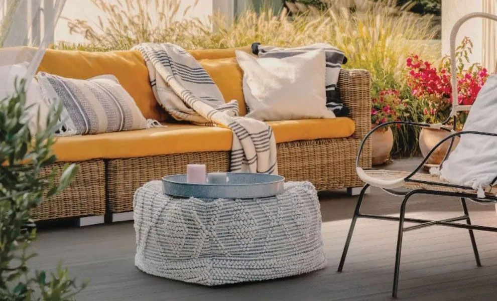 A patio scene featuring a wicker sofa with yellow cushions, a white knit ottoman, and a metal chair on a wooden deck.