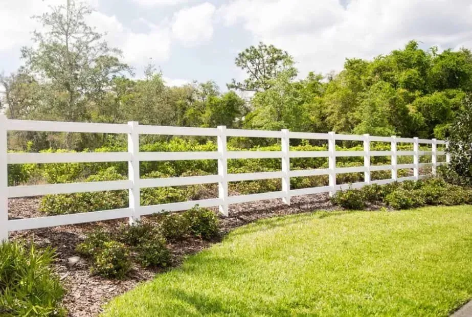 A white four-rail fence borders a manicured lawn and a garden bed, with dense trees and a bright sky in the background.