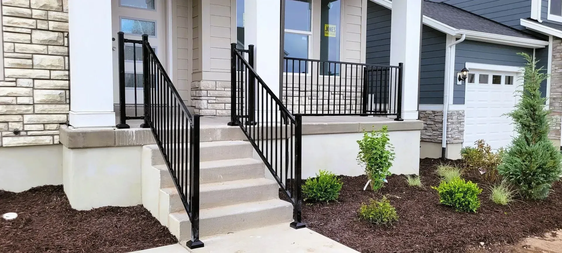 Concrete porch steps leading to a front door with black metal railings in front of a house with stone and siding facade.