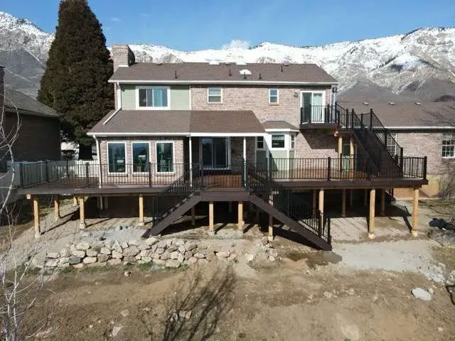 A two-story house with a large wooden deck, stairs, and brick and siding exterior, backed by snow-capped mountains.