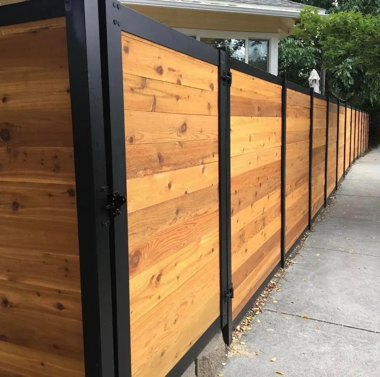 Modern fence featuring horizontal cedar wood planks framed in black metal posts, lining a sidewalk next to a house.