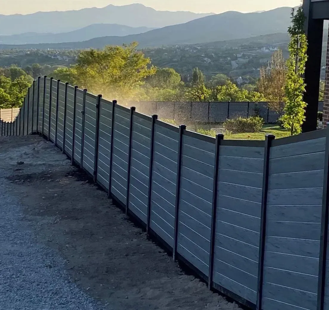 A long, modern gray privacy fence with black posts installed along a dirt path, overlooking mountains at sunset.