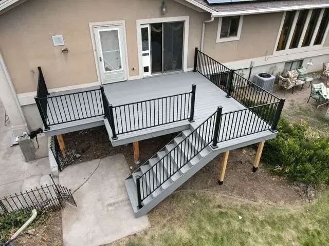 A high-angle view of a grey elevated backyard deck with black railings and stairs leading to a concrete patio.