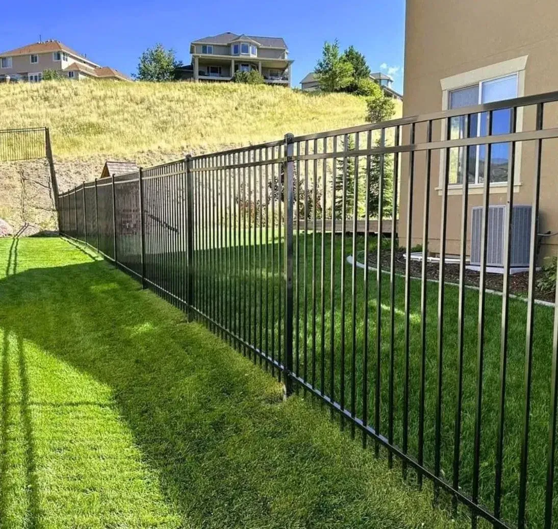 A black metal fence runs along a green lawn beside a beige house, with a grassy hill and neighboring homes in the back.