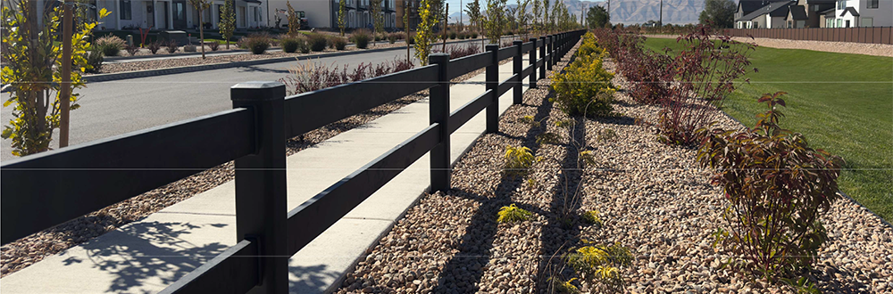 Black fence borders a gravel path beside landscaped plants on a sunny waterfront promenade.