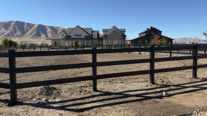 Black wooden fence in a dry field with houses and mountains under a clear blue sky